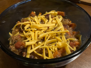 Blue glass bowl of chili on a wooden table. Cheddar cheese is sprinkled on top.
